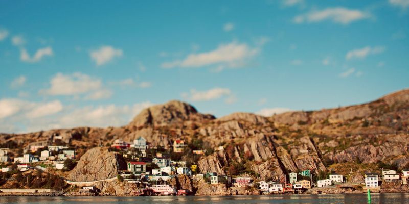 A photo taken from the ocean looking a scene featuring colourful houses, a rocky cliff, ocean and blue sky. The area is known as The Battery in the City of St. John's.